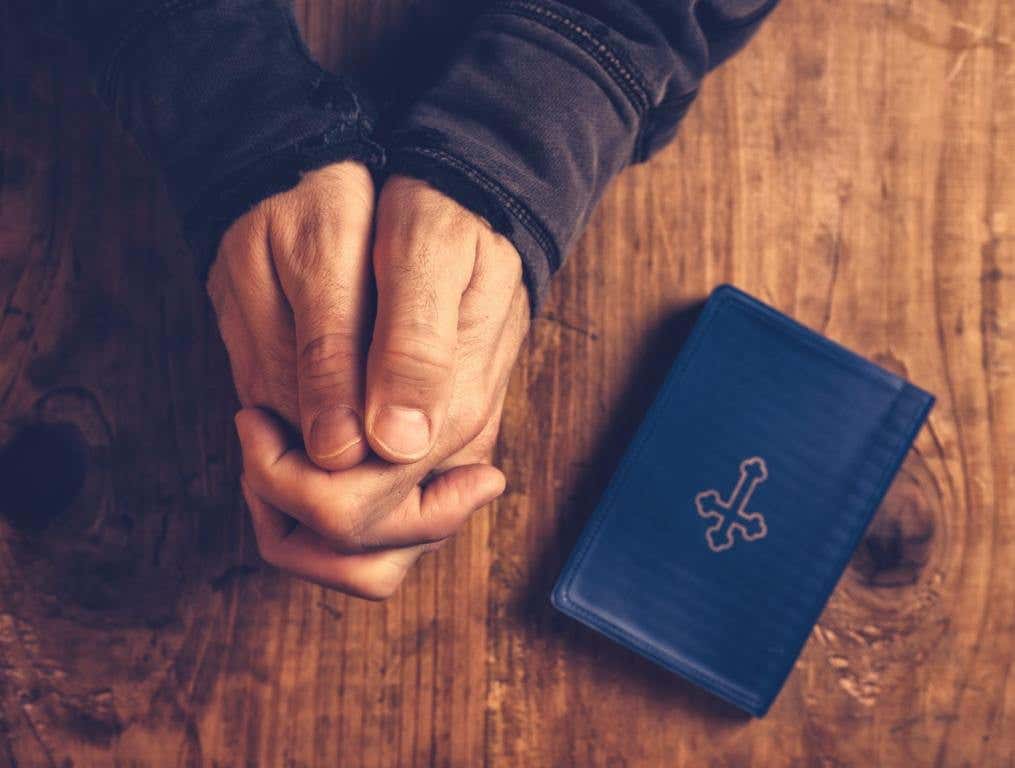 Christian man praying with hands folded and fingers crossed with Holy Bible by his side on wooden desk in church, top view