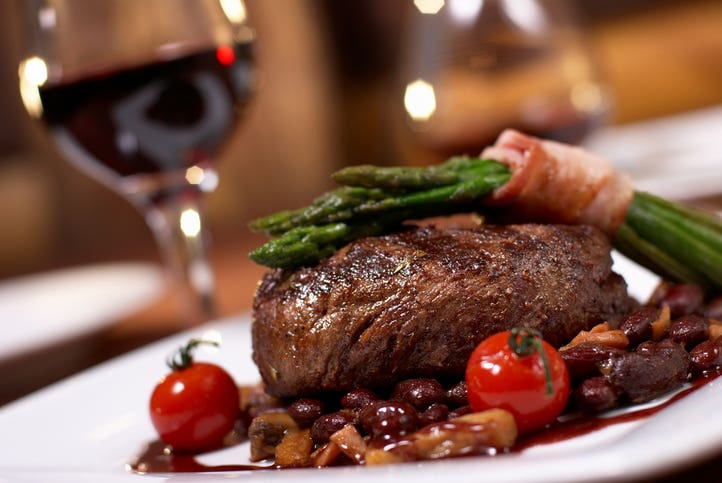 grilled beef with tomato served on a white plate with a glass of red wine behind it