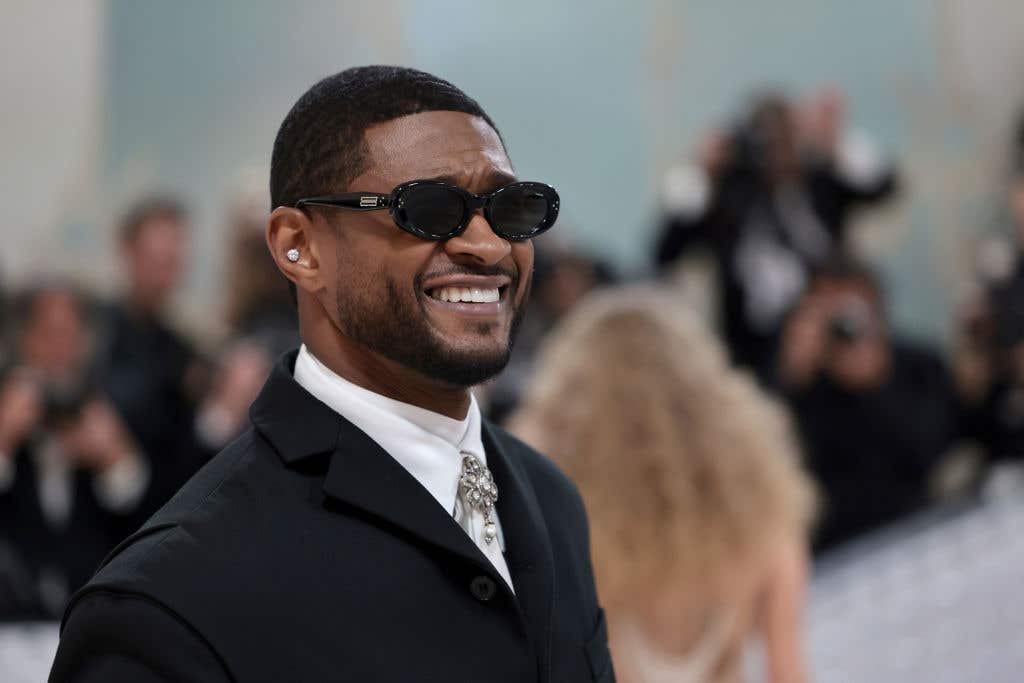 Usher wearing a suit and sunglasses on the red carpet of a the 2023 Met Gala event. He's smiling and he'll be performing at the Super Bowl.