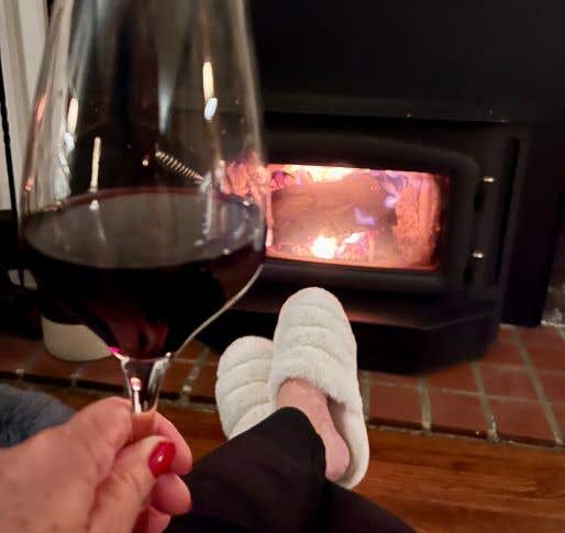 Feet wearing white slippers propped on a foot stool in front of a fire with a glass of wine in hand drinking Wines To Keep You Warm