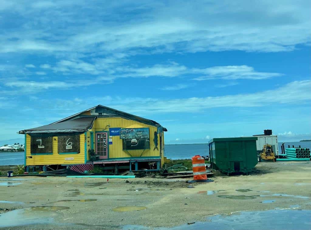 Single uninhabitated, yellow house sitting on sand with a green dumpster beside it in Matlacha