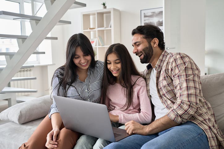 Happy indian family with child daughter having fun using laptop computer at home. Smiling parents and teen kid daughter laughing looking at device browsing or watching funny videos sitting on sofa.