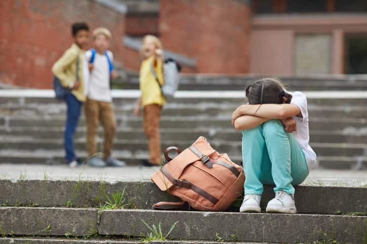 crying schoolgirl sitting on stairs outdoors with group of teasing children bullying her in background,