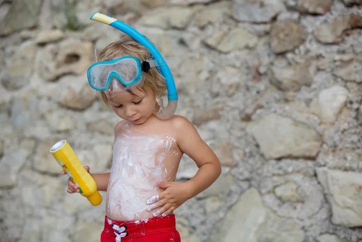 Little toddler child, holding sun cream, applying it on his body, sun protection during summer vacation.  Florida resorts with waterparks